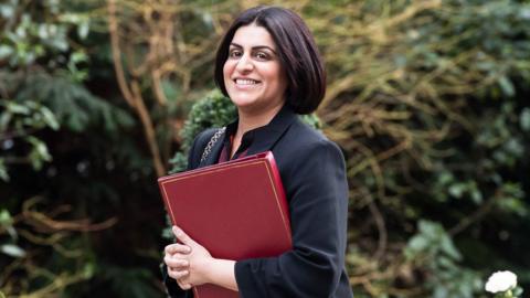 Home Secretary Shabana Mahmood walks outside of Downing Street. She wears a high-necked black jacket, black bobbed hair, and holds a burgundy folder.