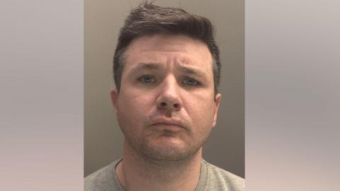 A police mugshot of Stephen Bates, 42, who has brown hair and is wearing a grey t-shirt, and is looking into the camera with a concerned expression.