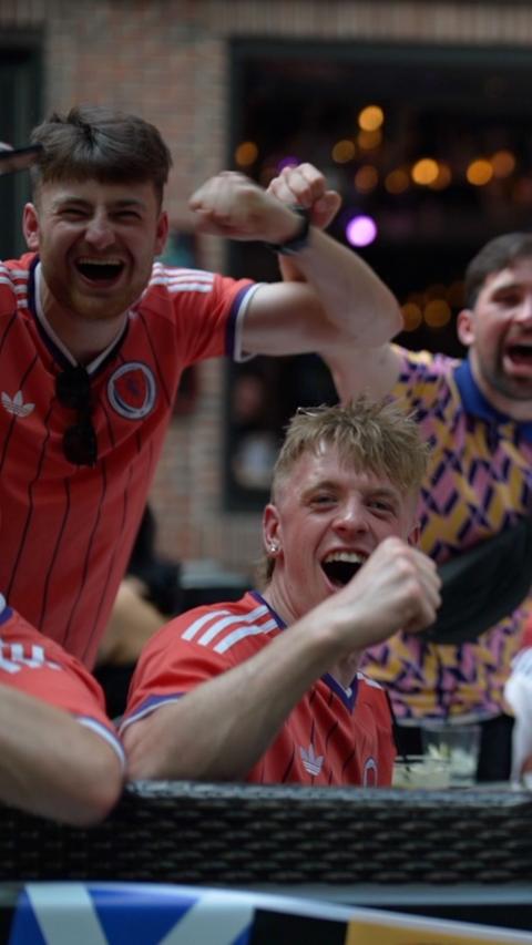 Two men in red shirts smile and raise their fists at the camera. One has brown hair and the other has blonde hair. Another man with brown hair is seen in the background wearing a pink, yellow and blue shirt.