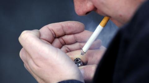 Close-up of a man in a black coat cupping a lighter in his hands as lights up a cigarette 