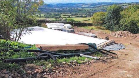 A hillside construction site with a partially built platform covered in white sheeting, scattered pipes and materials in the foreground, and a wide valley of fields and rolling hills stretching out under a partly cloudy sky.