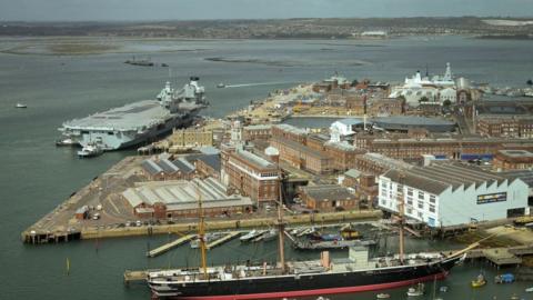 An aerial shot of Portsmouth Naval Base and the Historic Dockyard, showing the historical ship HMS Victory in the foreground and an aircraft carrier in the background