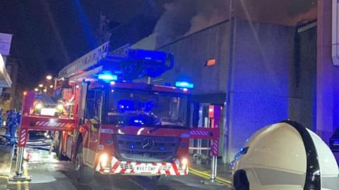 A fire engine with its lights on and two supports anchoring it on either side is parked in the street, with hoses seen trailing on the floor and firefighters in the distance. A concrete building behind the vehicle has smoke pouring from the roof and an orange glow, presumably from flames, appears through one of the windows. 