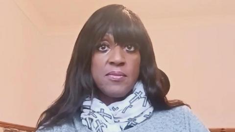 Grace Gomez, a black woman with long dark hair, is pictured facing the camera. She is wearing a white scarf with images of the Christian cross on it. 