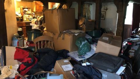 A kitchen and living room area filled with clutter. The floor cannot be seen and there are large boxes piled up among the clutter.