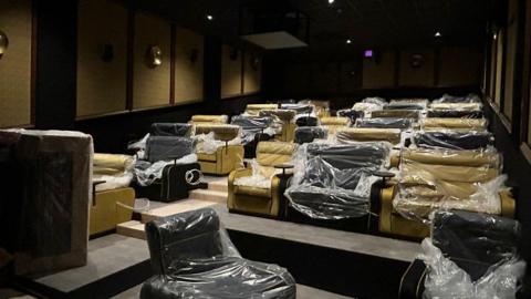 A small cinema with yellow and black plush armchairs, which are covered in plastic wrapping. The photo is taken from the end with the screen, looking up at the seats.