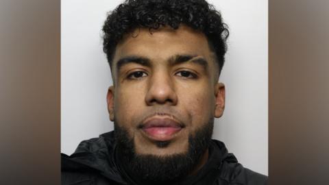A police mugshot of a man with curly black hair and a neat black beard. He is wearing a black coat