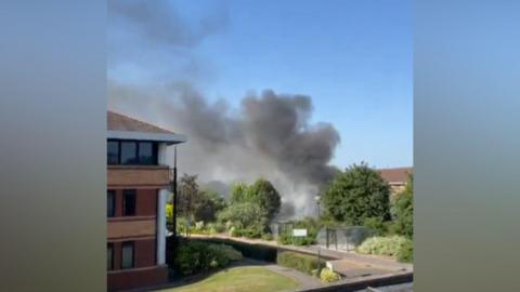 Smoke can be seen billowing from an area blocked by trees and shrubs. A large brick building is to the left along with a footpath.