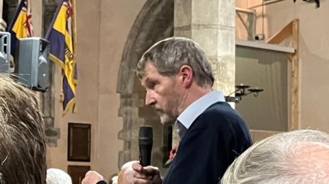 Simon Galton, from Harborough District Council, addresses a crowd, he is wearing a light blue collar shirt and black jumper with a poppy.