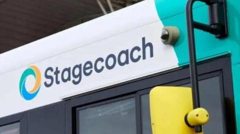 A picture of the side of a bus - which says the word stagecoach on it. It is a blue and white bus with a yellow wing mirror. There is a small camera next to the logo.