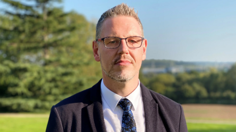 Picture of John Campion West Mercia Police and Crime Commissioner in a black suit with a flowery tie. He is wearing glasses and looking directly at the camera. There are trees and grass behind him