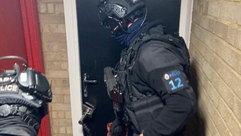 A police officer standing at a black door, preparing to force entry. He is wearing a black uniform and there is another police officer behind him.