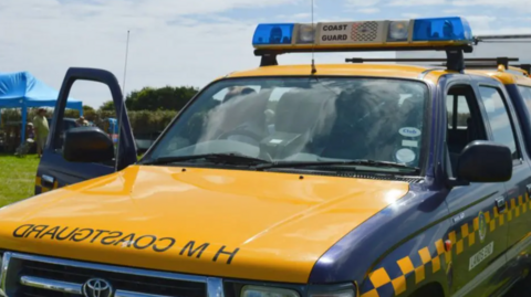 A Coastguard vehicle in yellow and dark blue is branded HM Coastguard on the bonnet, with a blue and white light set-up on the roof. 