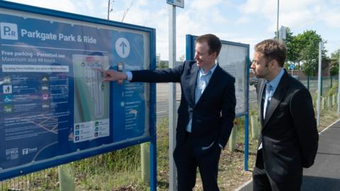 Two men in dark suits are outside, standing next to a large information board headed Parkgate Park & Ride. One of the men is pointing to a map on the board.
