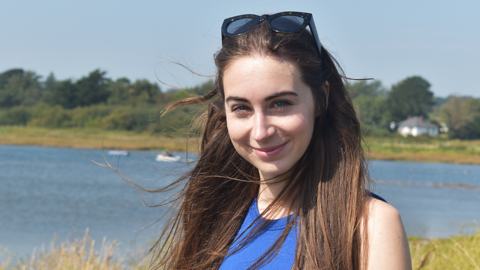 Bethany Handley is a white woman in her mid 20s. She has long light brown hair with sunglasses on top of her head in a head and shoulders shot. She wears a blue vest and is smiling at the camera. She is outdoors in front of a lake which is out of focus in the background surrounded by grass and trees.
