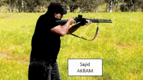 A man dressed in black and standing in a field of lush grass aims a shotgun
