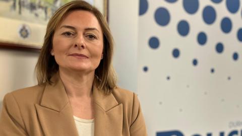 Durham Police Federation chair Louise Guest has a brunette bob hair cut and brown eyes. She is wearing a beige blaze with a creme top. She stands in front of a navy spotted sign which is blurred in the background.