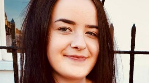 Emily Griffiths has long dark hair, and is sitting outside, with a building in the background. She has sunglasses on top of her head and is smiling at the camera.