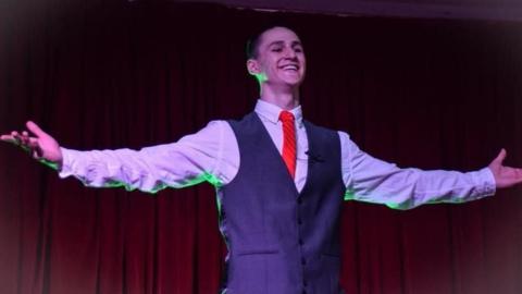 Conor is on stage, a red curtain behind him. He's wearing a tie and waistcoat and his hands are outstretched.