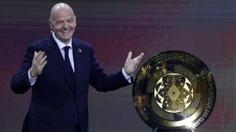 A smiling Gianni Infantino stands next to the Club World Cup trophy