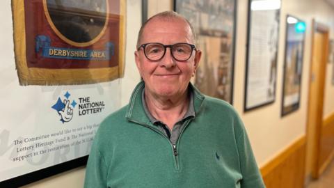 Alan Gascoyne standing in front of a picture which depicts the Shirebrook Colliery National Union of Mineworkers banner. He is wearing glasses and has a green jumper on.