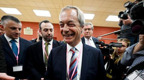 Nigel Farage grinning and surrounded by cameras. He's wearing a dark suit and a white shirt and a striped tie.