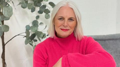 An woman with short white hair sits indoors on a grey cushioned seat, wearing a bright pink jumper and red trousers. A tall green houseplant in a woven basket stands behind them against a plain white wall.