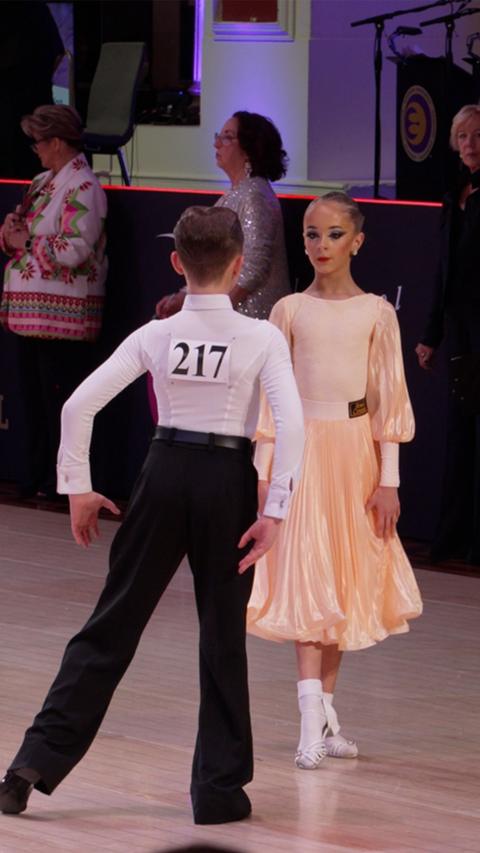 Two 11-year-old dancers on the ballroom floor. She is wearing a long light orange dress with white shoes and he is wearing black trousers and a white top. 