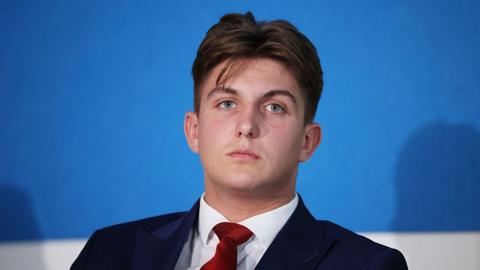 Reform UK councillor George Finch, a young man in a navy suit with a red tie, attends a press conference.