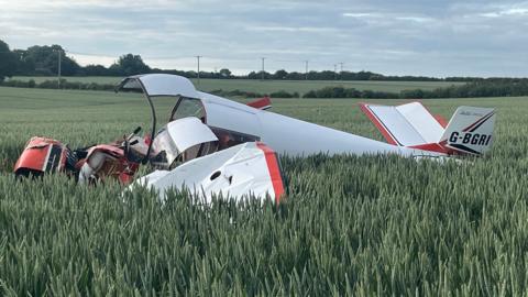 A close-up of the small plane in a field of wheat. It is mostly white livery, with a red strip, and the front of it is completely smashed. On the tail reads: "G-BGRI". In the background are hedges and small pylons, and the sun is starting to set.