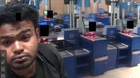 A CCTV image of a man in a supermarket with a row of self-service tills behind him. He has dark black hair and is wearing a dark green top