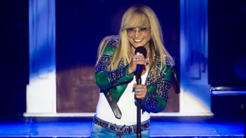 Anastacia performing in Milan - she has long blonde hair, glasses and a blue and green jacket