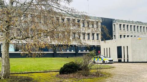 Police car outside a school building, with a tree in the foreground