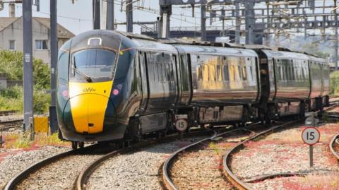 A Great Western Railway branded train is on a track leaving a train station. The train is a dark green with a yellow nose and the track below is pebbled,. There are some green bushes in the background and a beige building to the left of the image