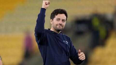 ryan mason pumps his right fist in the air and smiles whilst looking to his right. he wears a navy hoodie.