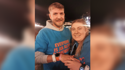 A man and a woman - Nick Bartrupe, and Amber Hird - stood in an embrace holding hands. Nick has short brown hair and is wearing a blue t-shirt with orange writing over a grey hoodie. He has a watch and a white wristband on his hand. Amber has long light brown hair and is wearing a blue hoodie. She has a ring on her finger. 