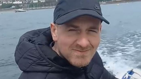 A man in a peaked cap and bomber jacket smiles while steeering a motor boat.