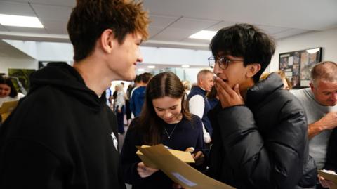 Two pupils smile and look shocked as they open brown envelopes, with another pupil reading their results on a piece of paper