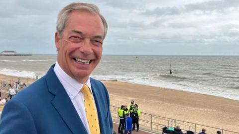 A smiling Nigel Farage wearing a light blue suit, with white shirt and yellow tie. He is stood above a sandy beach 