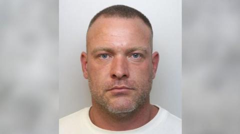 A mug shot of a man with grey hair in a white t-shirt