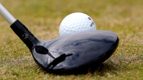A close-up of the head of a golf club beside a ball on a golf green.