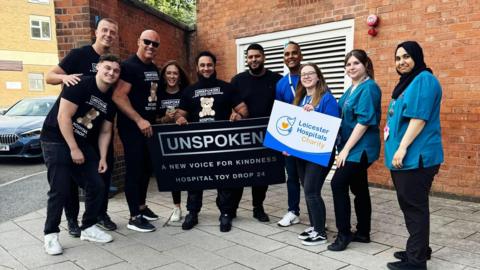 Atiq Rehman stands with six of his volunteers, who are dressed in black T-shirts with the Unspoken logo on. They are joined by four other people dressed in blue, and one holding a sign that reads "Leicester Hospitals Charity". 