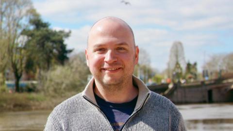 A close-up of a bald man smiling at the camera, wearing a sweater zipped up over a t-shirt. He stands in front of a river with trees and a bridge behind him.