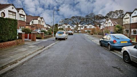 Residential properties on a cul-de-sac with cars parked on pavement. Their number plates are blocked out. It's a cloudy day.