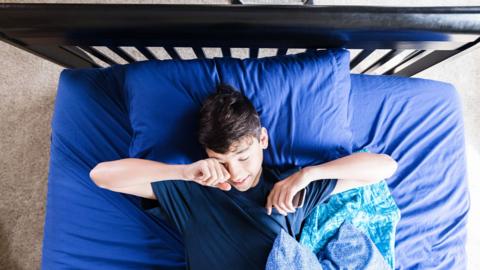 A teenage boy rubs his eyes after sleeping in on a Saturday morning.
