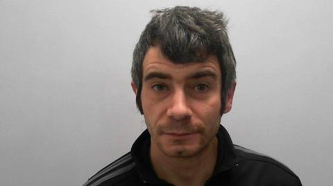 Police mugshot of Damian Hunter, a man with thick greying black hair, wearing a black tracksuit top.