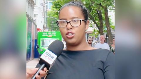 Woman stands on street and talks into BBC microphone that is being held in front of her
