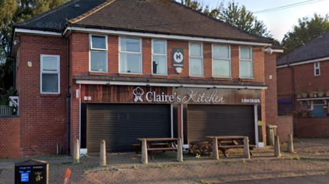 A cafe frontage with closed shutters. It is on a semi-residential street. There are wooden picnic benches and concrete posts outside