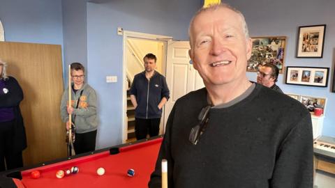 Steve Davis. He has short white hair and is wearing a black jumper with some glasses hooked on the neck. He is standing in front of a pool table and is looking directly at the camera and smiling.