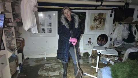 A woman wearing a dark blue padded coat and wellington boots carrying a shovel while standing inside a flood-damaged photography studio